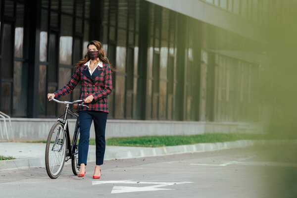 selective focus of businesswoman in plaid mask walking with bicycle 