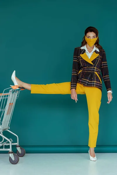 stylish woman in yellow mask putting leg on empty shopping cart on blue