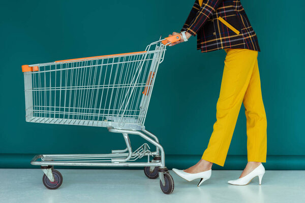 cropped view of trendy woman in yellow mask walking with empty shopping cart on blue