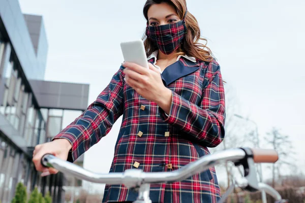 selective focus of businesswoman in plaid mask using smartphone near bicycle