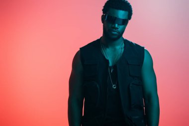Yakışıklı, modaya uygun, Afro-Amerikalı, fütüristik güneş gözlüklü, mavi ışıkta pembe poz veren adam. 