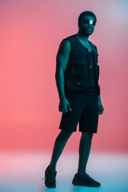Fütürist güneş gözlüklü, mavi ışıkta pembe poz veren yakışıklı Afro-Amerikalı adam. 