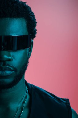 fashionable african american man in futuristic sunglasses posing on pink in blue light  clipart