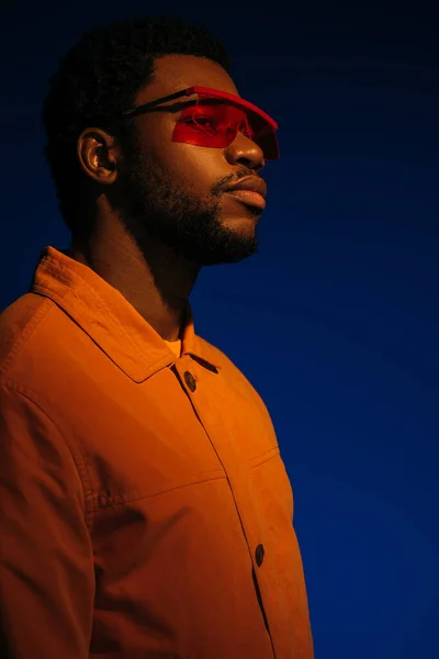 未来的な外観のファッショナブルなアフリカ系アメリカ人男性モデルと赤い光の中で青い上にポーズサングラス — ストック写真