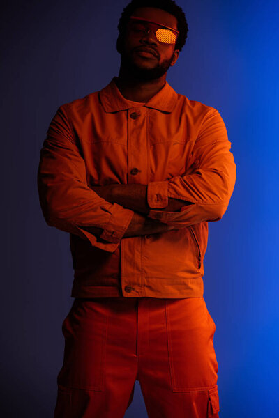 красивый африканский американец в футуристическом виде и солнечных очках, позирующих со скрещенными руками на синем в красном свете
 