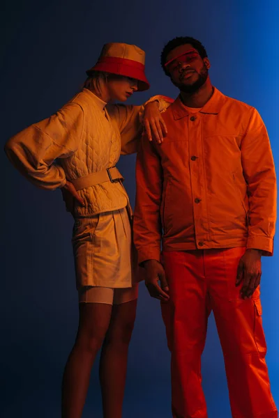 Interraciale Paar Futuristische Kleding Zonnebril Hoed Poseren Blauw Oranje Licht — Stockfoto