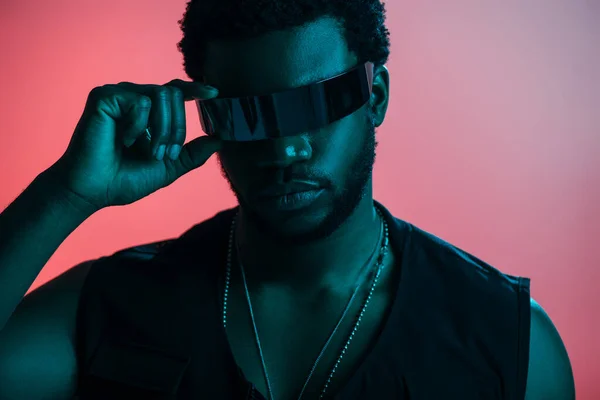 fashionable african american futuristic man in sunglasses posing on pink in blue light