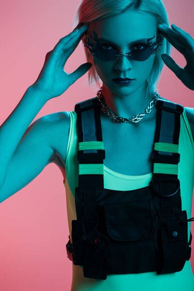 young fashionable model in futuristic bodysuit and fire-shaped sunglasses posing on pink in blue light 