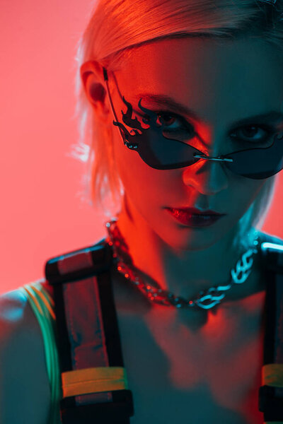 attractive futuristic girl posing in fire-shaped sunglasses on pink in red light