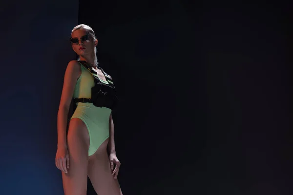 beautiful fashionable woman in futuristic bodysuit and fire-shaped sunglasses posing on dark