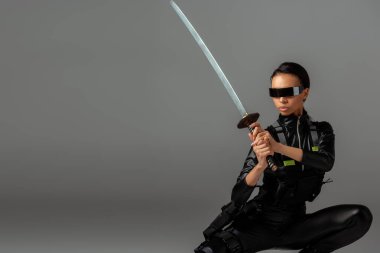 attractive futuristic african american woman in glasses sitting with sword on grey clipart