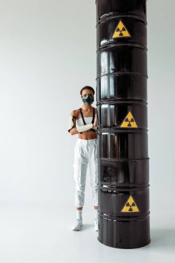 futuristic african american woman in safety mask near radioactive waste barrels on white background clipart