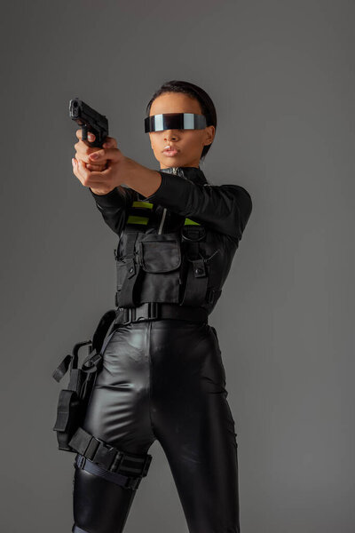 attractive futuristic african american woman in glasses aiming gun isolated on grey