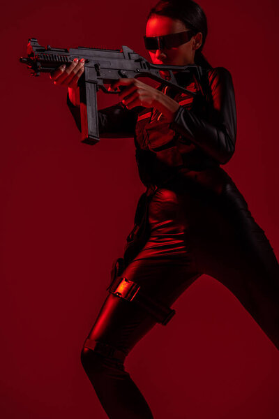 futuristic african american woman in glasses aiming assault rifle on red background