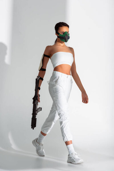 futuristic african american woman in safety mask walking with assault rifle on white background