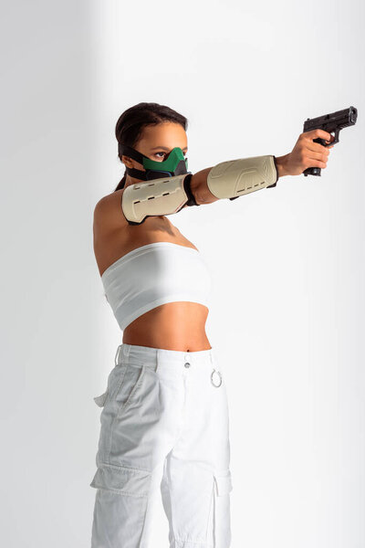 futuristic african american woman in safety mask aiming gun on white background