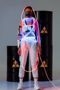futuristic african american woman in safety mask and neon light near radioactive waste barrels clipart