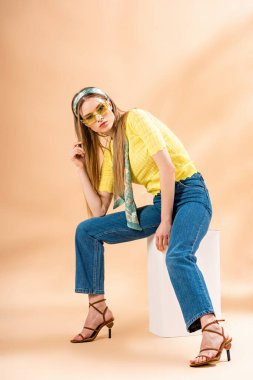 Kot pantolonlu, sarı tişörtlü, güneş gözlüklü, topuklu sandaletli ve bej üzerine oturan ipek eşarplı çekici bir kız.