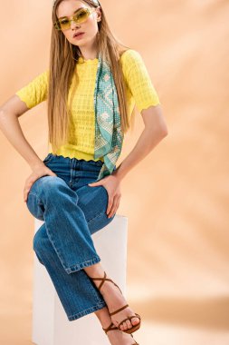 Kot pantolonlu güzel sarışın kız, sarı tişört, güneş gözlüğü, topuklu sandaletler ve bej üzerine oturan ipek eşarp.