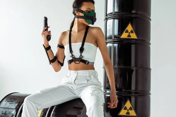 白い背景に放射性廃棄物の樽の近くに銃を持つ安全マスクの未来的なアフリカ系アメリカ人女性 — ストック写真