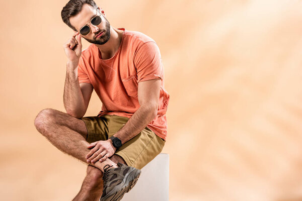 fashionable handsome young man in shorts, summer t-shirt and sunglasses sitting on white cube on beige