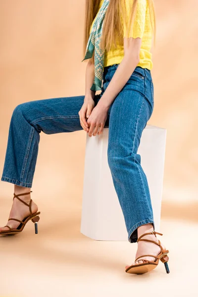 cropped view of girl sitting in jeans, yellow t-shirt, heeled sandals and silk scarf on beige