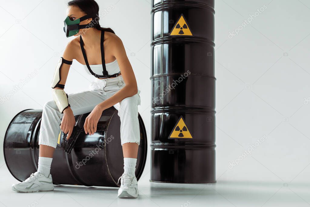 futuristic african american woman in safety mask with gun near radioactive waste barrels on white background