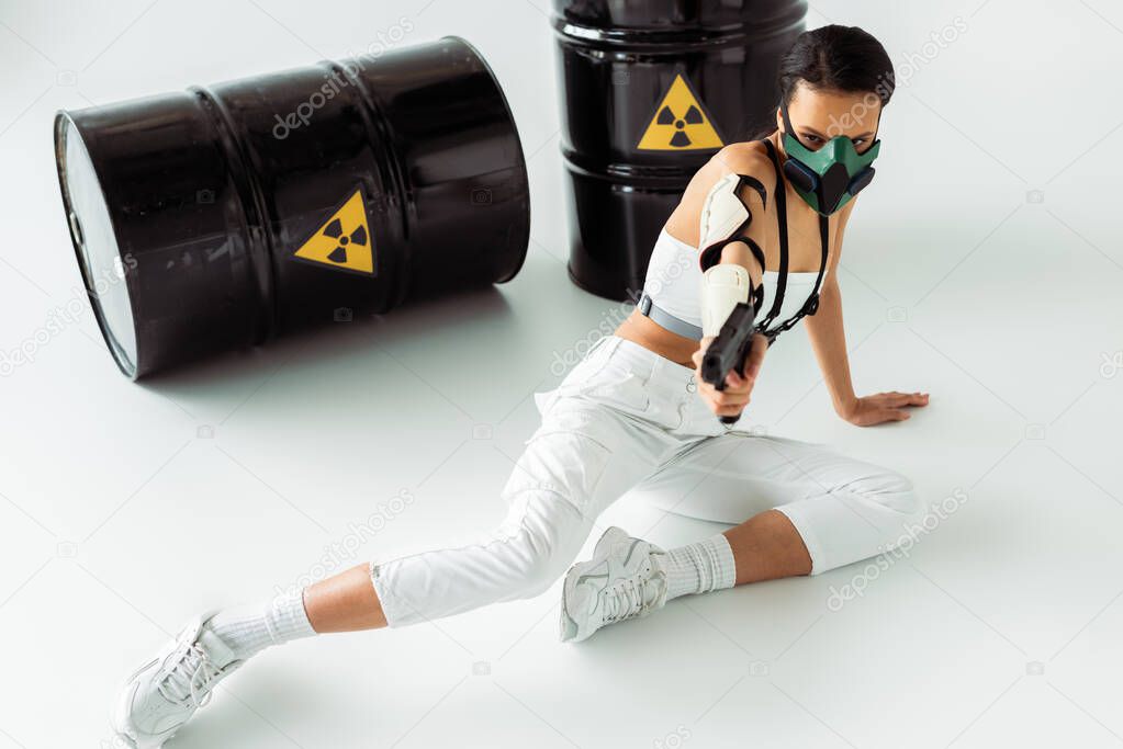 futuristic african american woman in safety mask aiming gun near radioactive waste barrels on white background