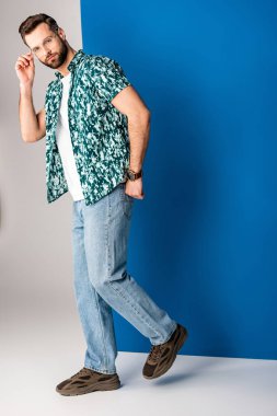 Yakışıklı sakallı adam gri ve mavi güneş gözlükleriyle yaz kıyafetleri içinde poz veriyor.