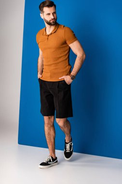 Sakallı yakışıklı adam şort ve kahverengi polo ile gri ve mavi üzerine poz veriyor. 