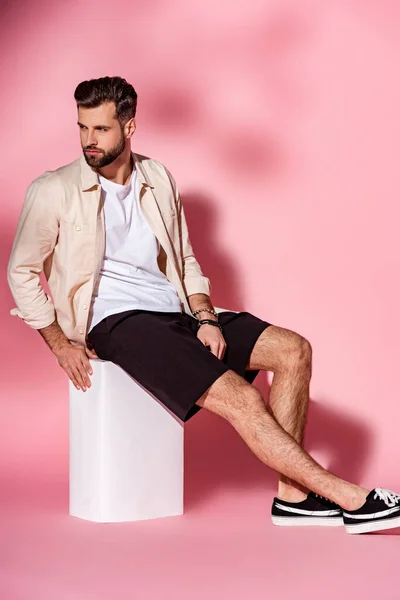 穿着夏装和短裤的英俊大胡子男人坐在粉色的白色立方体上 — 图库照片