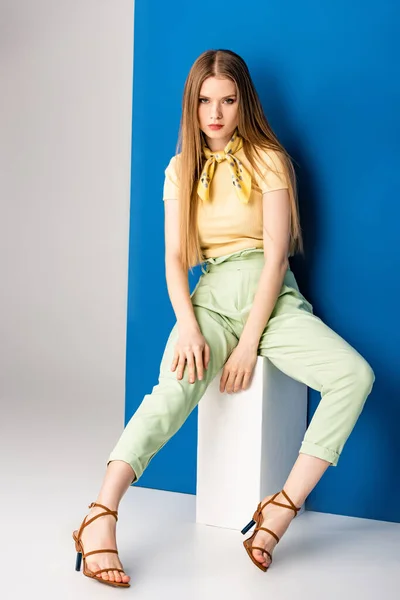 fashionable girl in green summer trousers and heeled sandals sitting on white cube on grey and blue