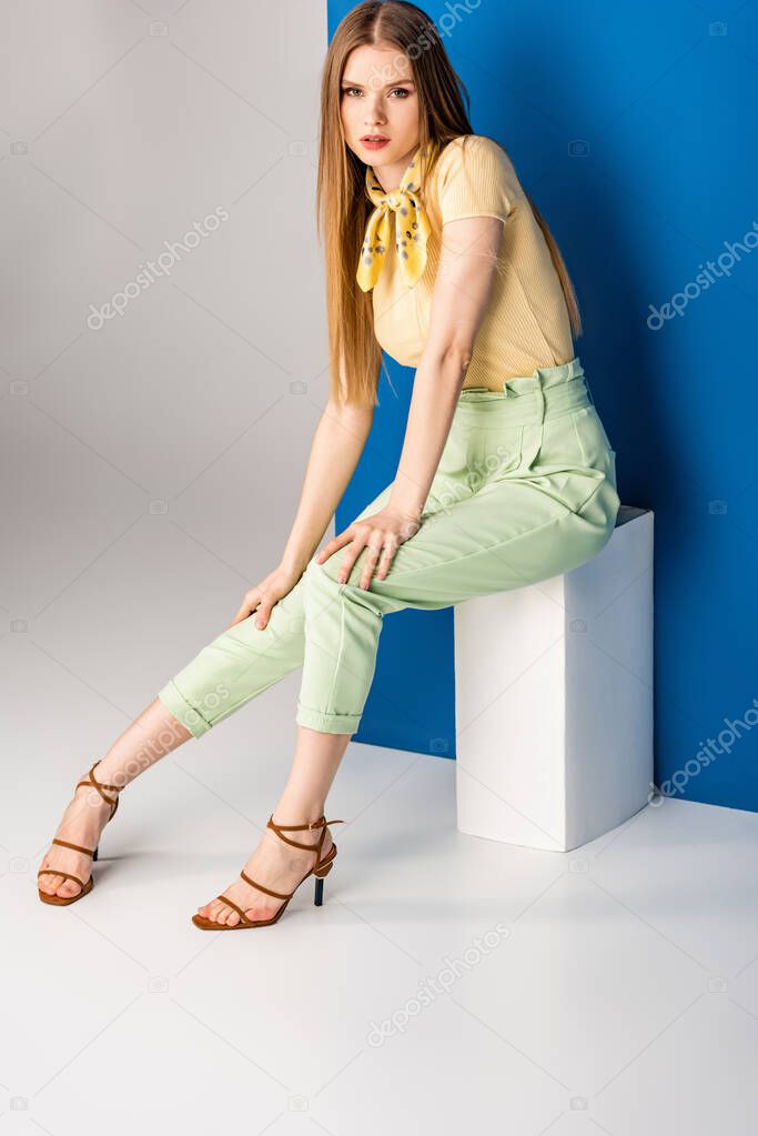 beautiful woman in green summer trousers and heeled sandals sitting on white cube on grey and blue