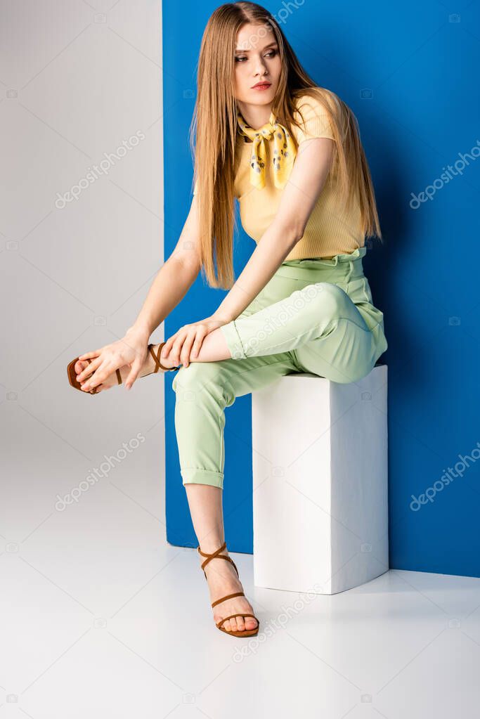 fashionable woman in green summer trousers and heeled sandals sitting on white cube on grey and blue