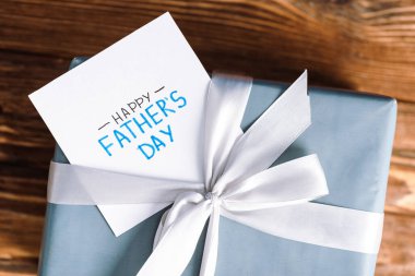 Mutlu Babalar Günü yazılı tebrik kartı manzarası ve tahta arka planda beyaz fiyonklu hediye kutusu.
