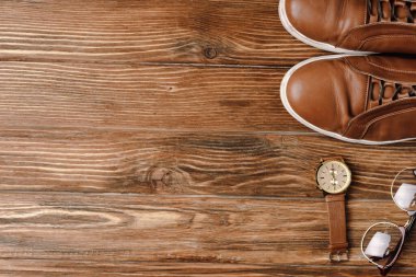 Erkeklerin kahverengi günlük ayakkabıları, kol saati ve tahta arka plandaki gözlüklerinin üst görüntüsü