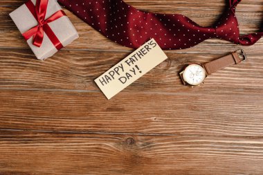 Kırmızı fiyonklu hediye kutusunun üst görüntüsü, mutlu babalar günü ve tahta arka planda erkek kravatı ve kol saati olan tebrik kartı.