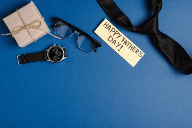Hediye kutusunun üst görüntüsü, mutlu babalar günü yazılı tebrik kartı ve erkek kravatı, kol saati ve mavi arka planda gözlük.