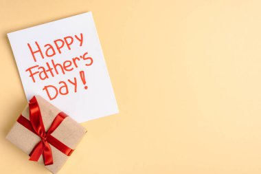 Kırmızı fiyonklu hediye kutusunun üst görüntüsü ve bej arka planda mutlu babalar günü yazılı tebrik kartı.