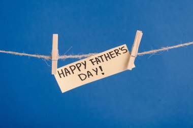 Beige paper greeting card with black lettering happy fathers day hanging on rope with clothespins isolated on blue clipart