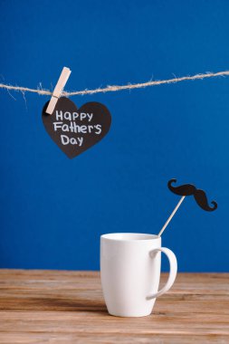 selective focus of white cup with decorative paper fake mustache and black greeting card with lettering happy fathers day isolated on blue clipart