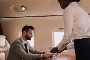 Afro-Amerikan hostes özel uçakta dizüstü bilgisayarda çalışan iş adamına bir fincan kahve veriyor.