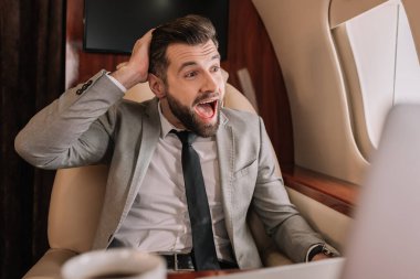 selective focus of shocked businessman with open mouth looking in porthole in private jet clipart