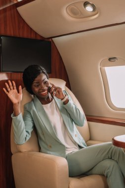 Mutlu Afro-Amerikan iş kadını özel uçakta akıllı telefonla konuşurken el sallıyor.
