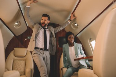 excited businessman screaming and touching ceiling while scared african american businesswoman sitting in private jet clipart