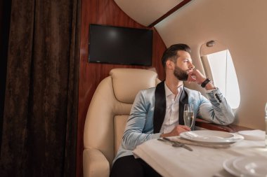 handsome, dreamy man looking in porthole while sitting at served table in private plane clipart