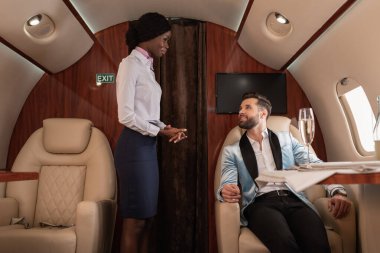 Çekici, gülümseyen Afro-Amerikan hostes yakışıklı, zarif bir adamın yanında duruyor özel jette servis masasında oturuyor.