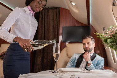 selective focus of attractive african american stewardess pouring champagne into glass near elegant man in private jet clipart