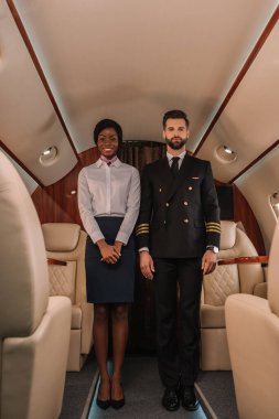 handsome, confident pilot and smiling african american stewardess looking at camera in private plane clipart