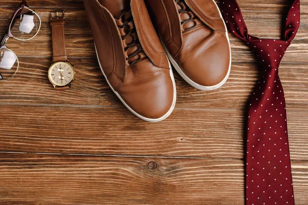 Top view of mens brown casual shoes, tie, wristwatch and glasses on wooden background